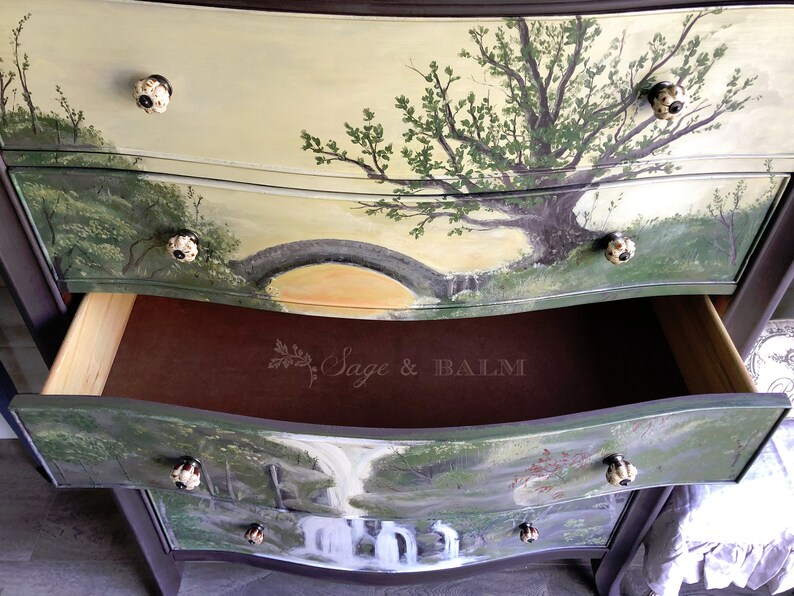 May include: A hand-painted dresser with three drawers features a nature scene on each drawer. The top drawer has a tree with a bridge in the background. The middle drawer is open and shows the brown interior. The bottom drawer has a waterfall scene. Each drawer has a unique knob.