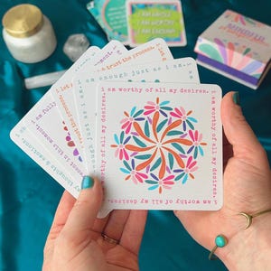 May include: A set of affirmation cards with colorful floral designs and positive messages. The cards feature phrases like "I am worthy of all my desires" and "I am enough just as I am".