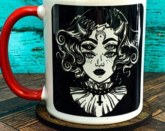 May include: A white ceramic mug with a red rim and handle. The mug features a black and white illustration of a woman with three eyes, horns, and a cross necklace. The background is black.