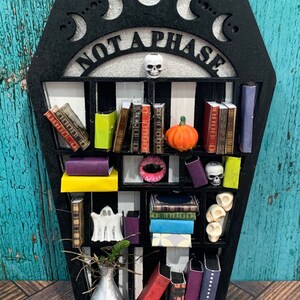 May include: A black coffin-shaped bookshelf with the words "Not a Phase" on the top. The bookshelf has multiple shelves with miniature books, skulls, a ghost, a pumpkin, and a vase with a plant.