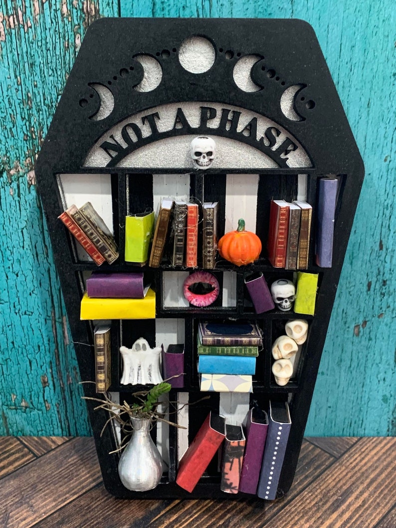 May include: A black coffin-shaped bookshelf with the words "Not a Phase" on the top. The bookshelf has multiple shelves with miniature books, skulls, a ghost, a pumpkin, and a vase with a plant.