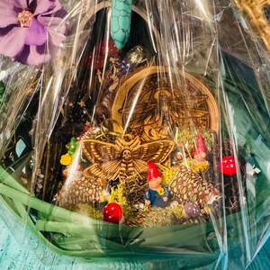Puede incluir: Una cesta de regalo decorativa llena de figuras en miniatura, incluyendo gnomos, setas y una polilla de madera. La cesta est&aacute; envuelta en celof&aacute;n transparente e incluye una flor morada y una varita de cristal turquesa.