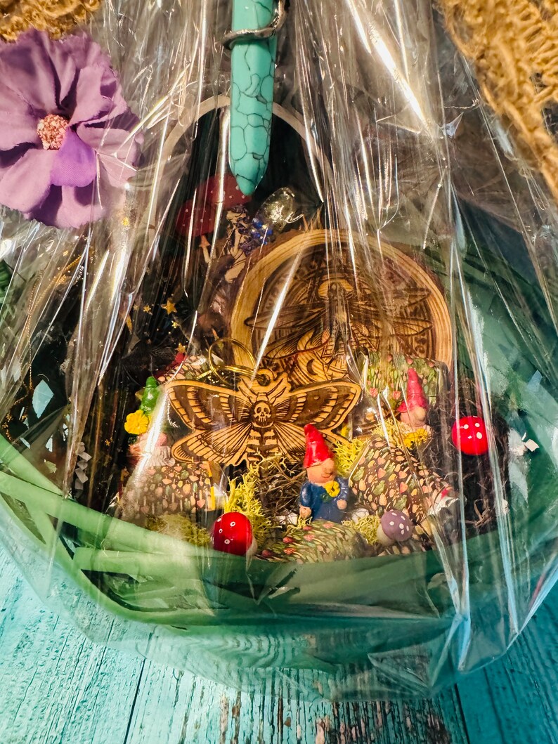 Puede incluir: Una cesta de regalo decorativa llena de figuras en miniatura, incluyendo gnomos, setas y una polilla de madera. La cesta est&aacute; envuelta en celof&aacute;n transparente e incluye una flor morada y una varita de cristal turquesa.