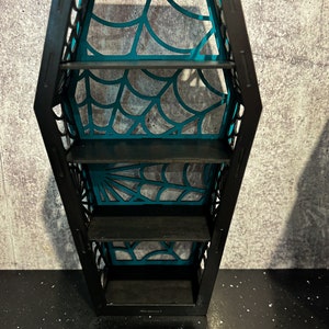 May include: A black coffin-shaped shelf with four shelves. The shelves are decorated with a teal spiderweb design.