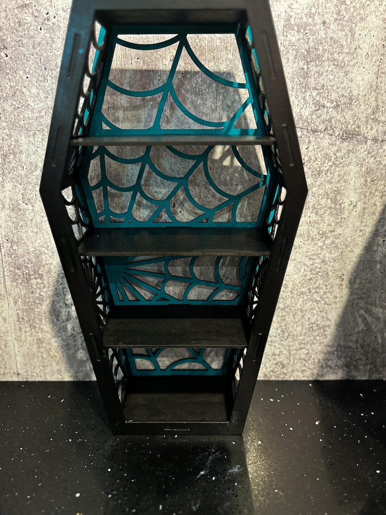 May include: A black coffin-shaped shelf with four shelves. The shelves are decorated with a teal spiderweb design.