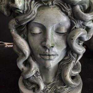 May include: A weathered, gray-green sculpture of Medusa's head with closed eyes and serpentine hair. The statue has a textured surface and is mounted on a circular base. The sculpture evokes a classical, mythological theme.