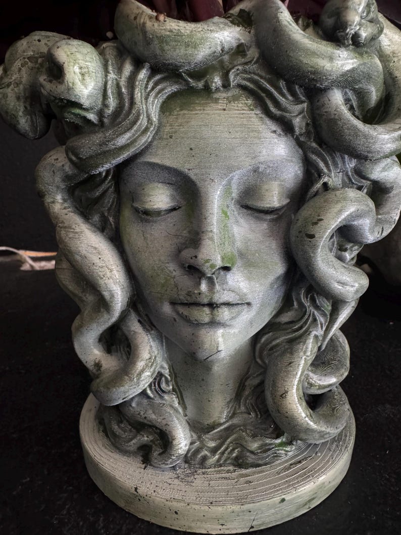 May include: A weathered, gray-green sculpture of Medusa's head with closed eyes and serpentine hair. The statue has a textured surface and is mounted on a circular base. The sculpture evokes a classical, mythological theme.