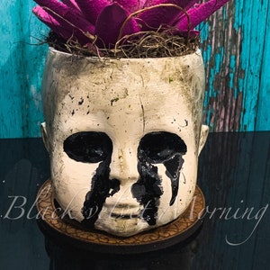May include: A unique, handcrafted planter shaped like a doll's head, painted in off-white with black accents. The doll's eyes are black, with black paint dripping down the face. The planter holds a vibrant, spiky, magenta succulent. The planter sits on a wooden coaster.