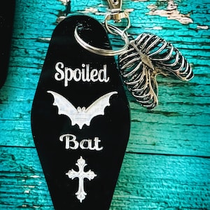 Puede incluir: Un llavero negro estilo motel con el texto grabado en blanco Spoiled Bat, una silueta de murci&eacute;lago y una cruz. Unido a la anilla de metal plateado hay un dije de metal plateado con forma de caja tor&aacute;cica humana y un cierre de mosquet&oacute;n.