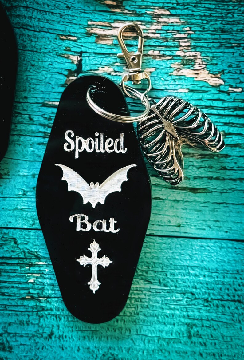 Puede incluir: Un llavero negro estilo motel con el texto grabado en blanco Spoiled Bat, una silueta de murci&eacute;lago y una cruz. Unido a la anilla de metal plateado hay un dije de metal plateado con forma de caja tor&aacute;cica humana y un cierre de mosquet&oacute;n.