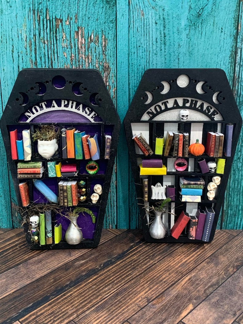 May include: Two black coffin-shaped shelves with the text "Not a Phase" on the front. Each shelf is decorated with books, skulls, a pumpkin, and other Halloween-themed items.