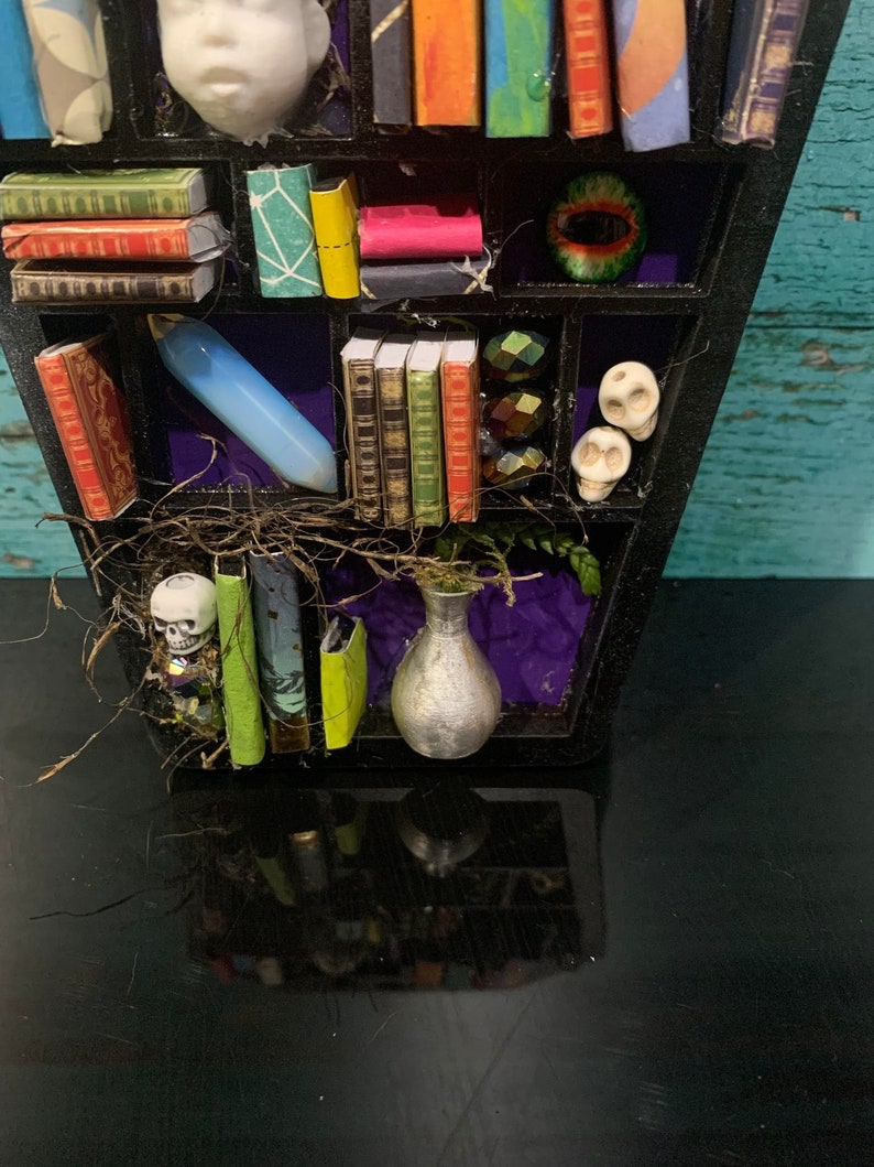 May include: A black coffin-shaped bookshelf with miniature books, skulls, a vase, and a glass eye. The bookshelf is decorated with purple and green accents.
