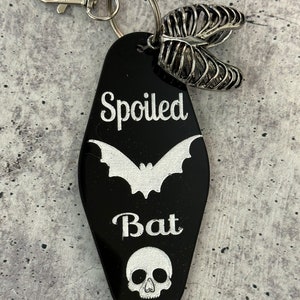 Puede incluir: Un llavero negro estilo motel con el texto grabado en blanco "Spoiled Bat", una silueta de murci&eacute;lago y un icono de calavera. El llavero incluye un cierre de mosquet&oacute;n plateado, una anilla y un dije plateado con forma de caja tor&aacute;cica.