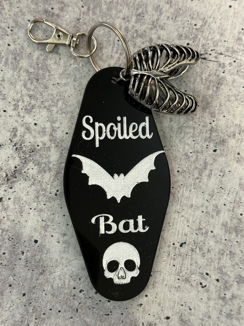 Puede incluir: Un llavero negro estilo motel con el texto grabado en blanco "Spoiled Bat", una silueta de murci&eacute;lago y un icono de calavera. El llavero incluye un cierre de mosquet&oacute;n plateado, una anilla y un dije plateado con forma de caja tor&aacute;cica.