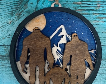 May include: A round wooden ornament with a black frame, featuring a silhouette of three Bigfoot figures standing in front of a mountain range. The background is a blue sky with white stars and a full moon.