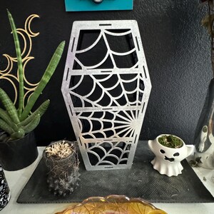 May include: A silver metal coffin-shaped decorative object with a spiderweb design. The object is sitting on a black surface.