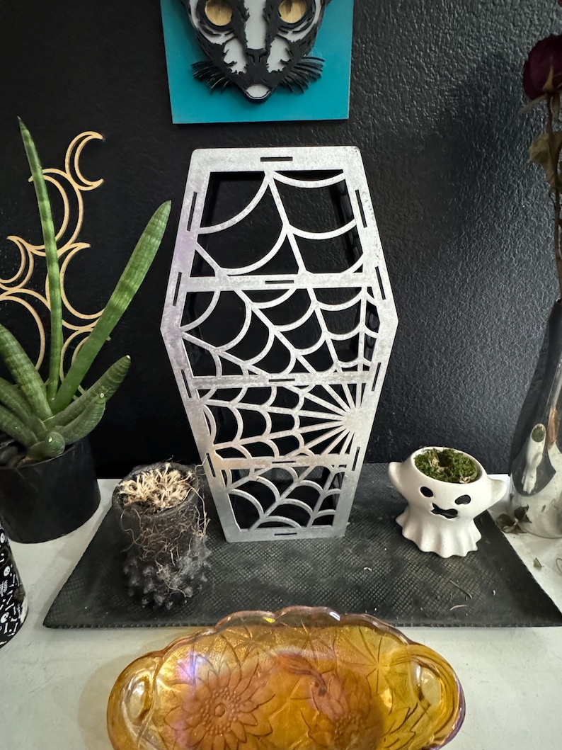 May include: A silver metal coffin-shaped decorative object with a spiderweb design. The object is sitting on a black surface.