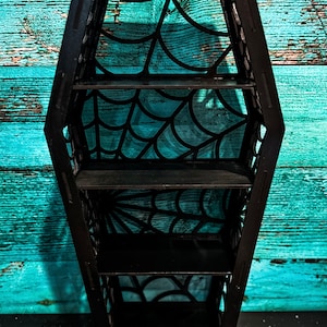 May include: Black coffin-shaped shelf with a spiderweb design. The shelf has four shelves.