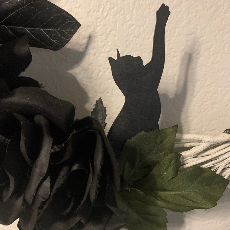 K&ouml;nnte beinhalten: Eine schwarze Katzensilhouette mit erhobenen Pfoten ist an einem wei&szlig;en Zweigkranz befestigt. Der Kranz ist mit schwarzen Samtrose und gr&uuml;nen Bl&auml;ttern verziert.