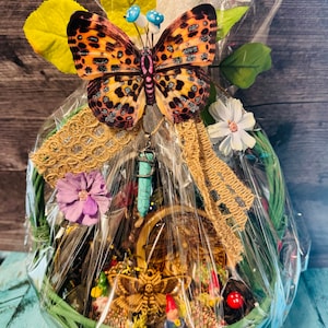 Puede incluir: Un juego de regalo de cesta de mimbre verde envuelto en celof&aacute;n, adornado con una gran mariposa, flores y un colgante turquesa. La cesta contiene una escena en miniatura con una polilla, gnomos y setas.