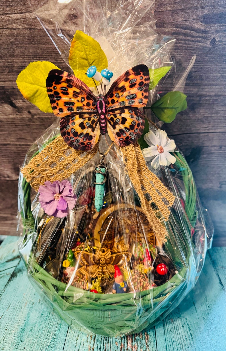 Puede incluir: Un juego de regalo de cesta de mimbre verde envuelto en celof&aacute;n, adornado con una gran mariposa, flores y un colgante turquesa. La cesta contiene una escena en miniatura con una polilla, gnomos y setas.