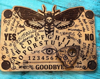 Könnte beinhalten: Ein Ouija-Brett aus Holz mit einem schwarz-weißen Mottiv. Das Brett enthält die Buchstaben A-Z, die Zahlen 1-9 und die Wörter "YES", "NO" und "GOODBYE". Ein schwarz-weißes Planchette ist enthalten.