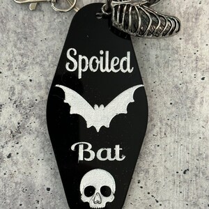 Puede incluir: Un llavero negro estilo motel con el texto blanco Spoiled Bat, un gr&aacute;fico de murci&eacute;lago blanco y un gr&aacute;fico de calavera blanco. El llavero incluye una anilla de metal plateado, un cierre de mosquet&oacute;n y un dije de caja tor&aacute;cica de metal plateado.