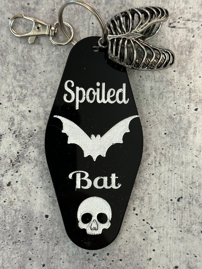 Puede incluir: Un llavero negro estilo motel con el texto blanco Spoiled Bat, un gr&aacute;fico de murci&eacute;lago blanco y un gr&aacute;fico de calavera blanco. El llavero incluye una anilla de metal plateado, un cierre de mosquet&oacute;n y un dije de caja tor&aacute;cica de metal plateado.