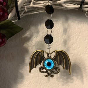 K&ouml;nnte beinhalten: Ein blaues Auge mit einer schwarzen Pupille ist von einem goldenen Metallrahmen mit einem Schlangenmuster umgeben. Das Auge ist an einem Paar goldener Fl&uuml;gel aus Metall befestigt und h&auml;ngt an einer schwarzen Perlenkette.