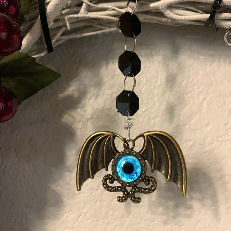 K&ouml;nnte beinhalten: Ein blaues Auge mit einer schwarzen Pupille ist von einem goldenen Metallrahmen mit einem Schlangenmuster umgeben. Das Auge ist an einem Paar goldener Fl&uuml;gel aus Metall befestigt und h&auml;ngt an einer schwarzen Perlenkette.