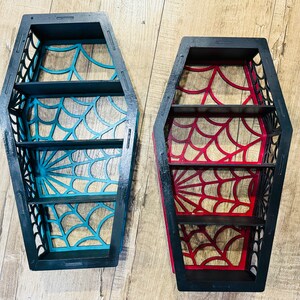 May include: Two black wooden coffin-shaped shelves with a spiderweb design. One shelf is painted teal blue and the other is painted red.