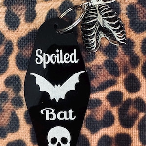 Puede incluir: Un llavero negro estilo motel con el texto blanco Spoiled Bat, un gr&aacute;fico de murci&eacute;lago blanco y un icono de calavera blanco. El llavero cuenta con un cierre de mosquet&oacute;n plateado y un dije de metal plateado con forma de caja tor&aacute;cica humana.