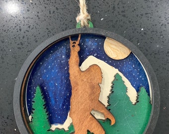 May include: A round wooden ornament with a silhouette of Bigfoot standing in a forest with a full moon in the background. The ornament is painted in shades of blue, green, brown, and white.