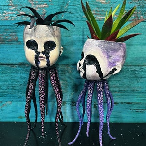 Puede incluir: Dos macetas de cerámica con forma de cabeza de muñeca con caras pintadas en blanco y negro y lágrimas pintadas en negro. Las macetas están sobre patas de pulpo negras con detalles en rosa y morado. Las macetas están llenas de suculentas verdes.