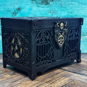 May include: A black rectangular wooden box featuring intricate laser-cut details, including a pentagram on the side and a gold-colored skull and pentagram emblem on the front latch. The box has arched window-like cutouts and ornate carvings.