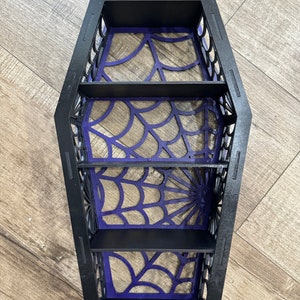 May include: Black wooden coffin-shaped shelf with four shelves. The shelves are decorated with a purple spiderweb design.