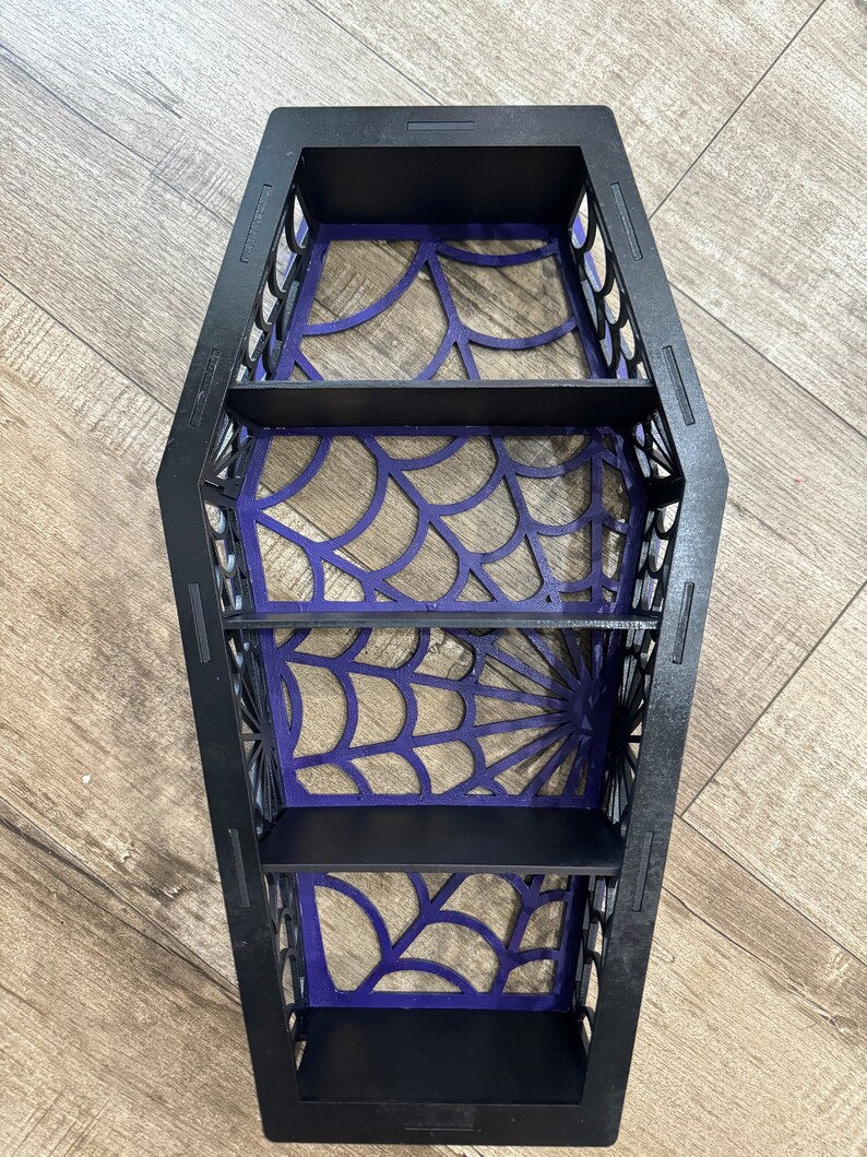 May include: Black wooden coffin-shaped shelf with four shelves. The shelves are decorated with a purple spiderweb design.