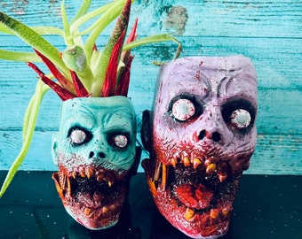 May include: Two zombie-themed planters, one with a green face and the other with a purple face. Both planters have red and white eyes and open mouths with teeth.