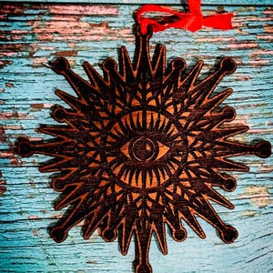May include: A wooden ornament with a sunburst design featuring an eye in the center. The ornament is brown with intricate cut-out details and a red ribbon for hanging. The background is a distressed blue wooden surface.