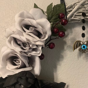 K&ouml;nnte beinhalten: Ein grauer und schwarzer Halloween-Kranz mit drei grauen Rosen, roten Beeren und einem h&auml;ngenden Fledermaus-Anh&auml;nger mit einem blauen Auge.