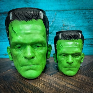 May include: Two green Frankenstein-themed planters, one large and one small, with black hair and facial features. The planters are made of a smooth, green material and have a realistic appearance.