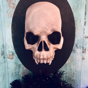 Op de afbeelding: Een schedelvormig ornament met een lichtbeige afwerking, gemonteerd op een zwarte ovale basis. De schedel heeft gedetailleerde kenmerken en staat tegen een turquoise en houtnerf achtergrond. Het ornament maakt deel uit van een Halloween- of gothic-thema decoratie.