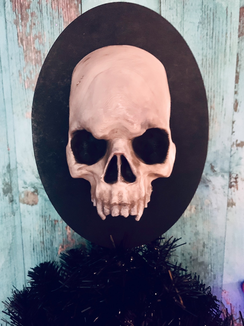 Op de afbeelding: Een schedelvormig ornament met een lichtbeige afwerking, gemonteerd op een zwarte ovale basis. De schedel heeft gedetailleerde kenmerken en staat tegen een turquoise en houtnerf achtergrond. Het ornament maakt deel uit van een Halloween- of gothic-thema decoratie.