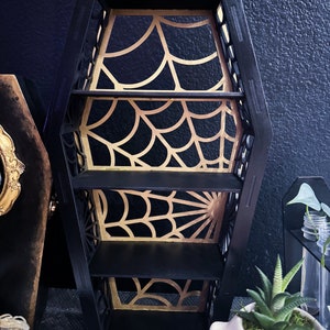 May include: Black coffin-shaped shelf with three shelves. The shelves are decorated with a gold spiderweb design.