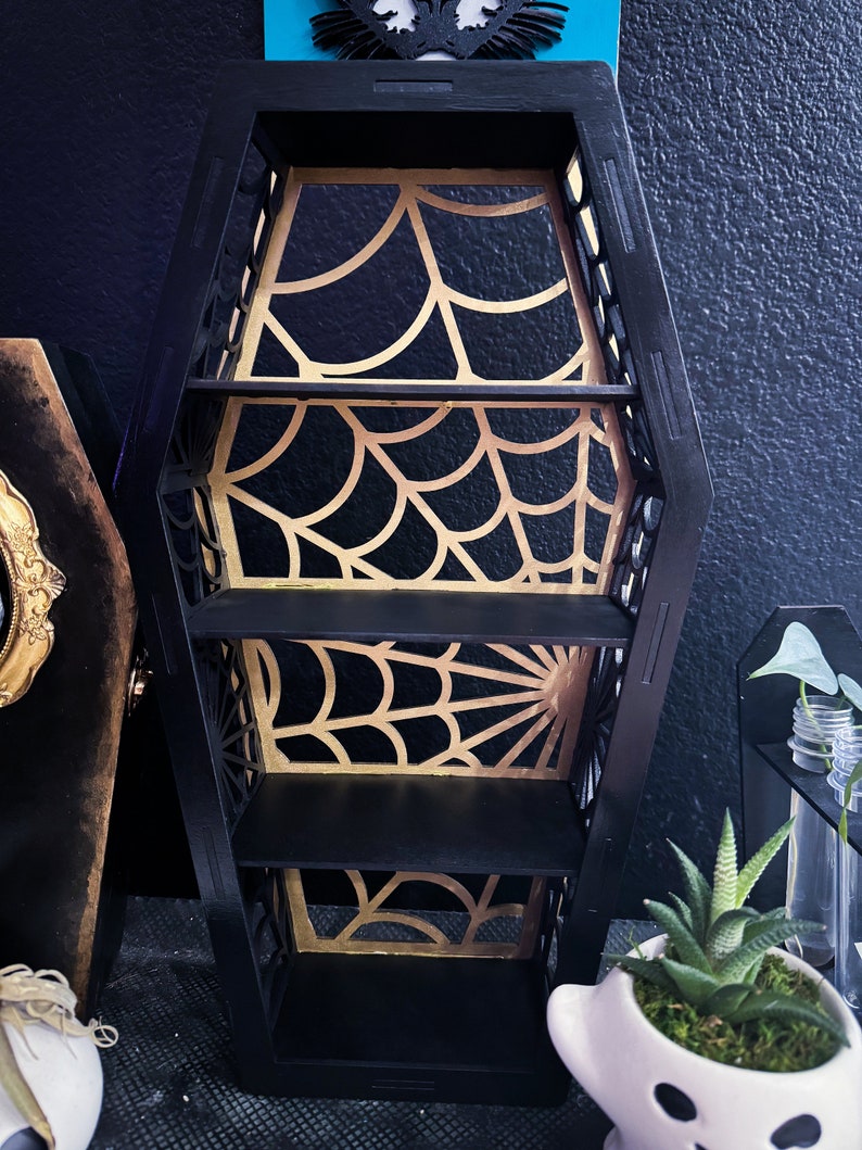 May include: Black coffin-shaped shelf with three shelves. The shelves are decorated with a gold spiderweb design.