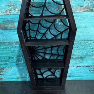 May include: Black wooden coffin-shaped shelf with four shelves. The shelves are decorated with a spiderweb design.