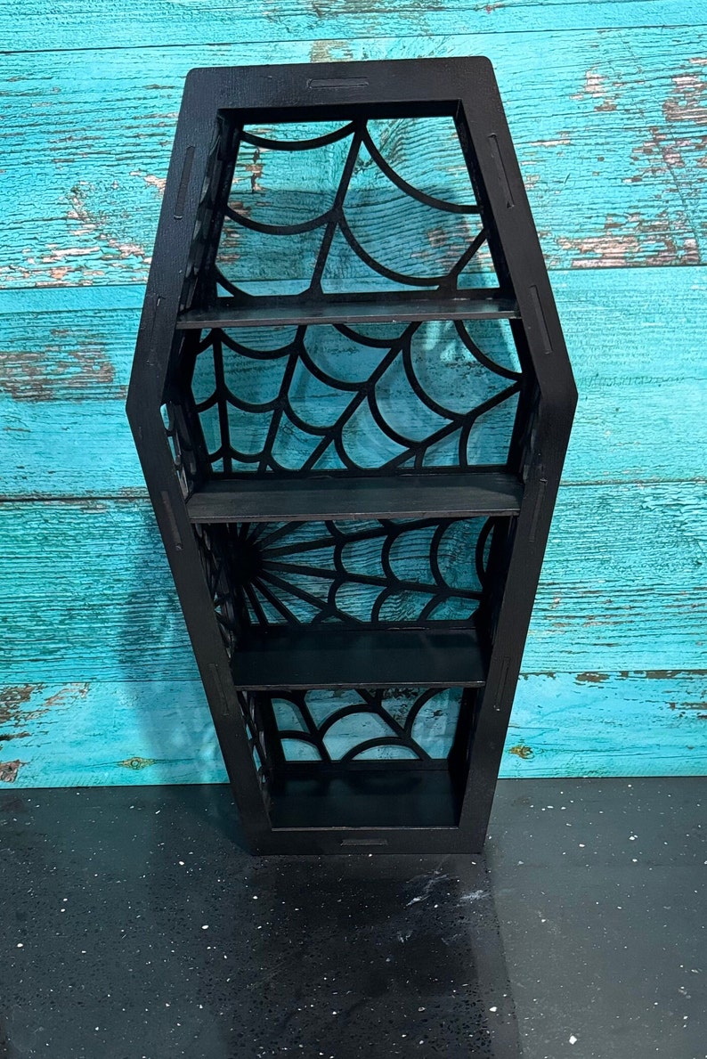 May include: Black wooden coffin-shaped shelf with four shelves. The shelves are decorated with a spiderweb design.