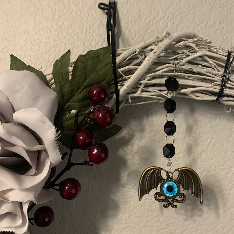 K&ouml;nnte beinhalten: Ein wei&szlig;er Zweigkranz mit einer grauen Stoffrose, roten Beeren und einem h&auml;ngenden Ornament. Das Ornament ist eine goldene Fledermaus mit einem blauen Auge und einer schwarzen Perlenkette.