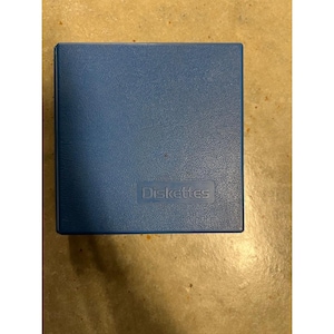 Peut inclure: Un étui carré bleu avec le mot "Diskettes" imprimé sur le devant. L'étui a une surface texturée et une forme rectangulaire. L'étui est probablement conçu pour contenir des disquettes.
