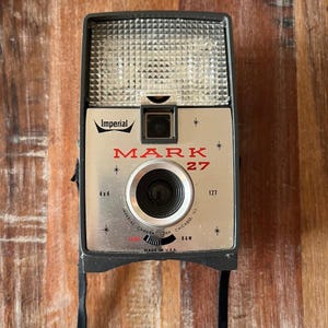 May include: A vintage Imperial Mark 27 camera with a silver faceplate, black body, and a top-mounted flash unit. The front features the text Imperial, MARK 27, 4x4, 127, and Imperial Camera Corp, Chicago, Ill, Made in U.S.A. with a black strap.